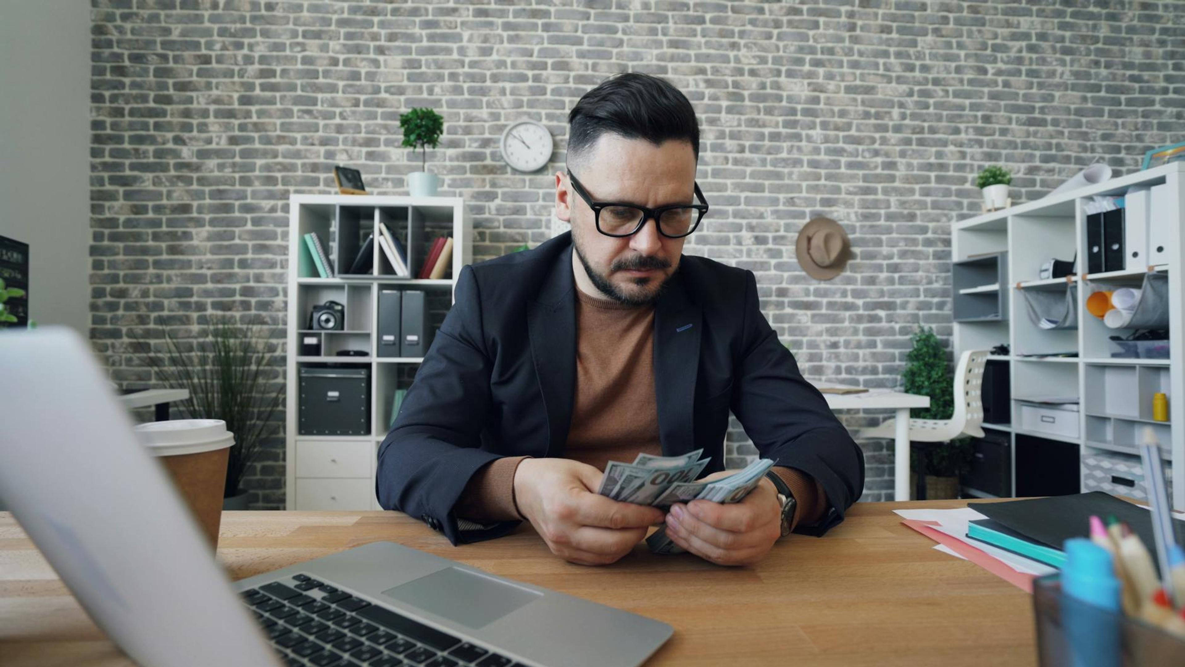 Businessman counting cash at a desk in a modern office setting with a laptop and documents.