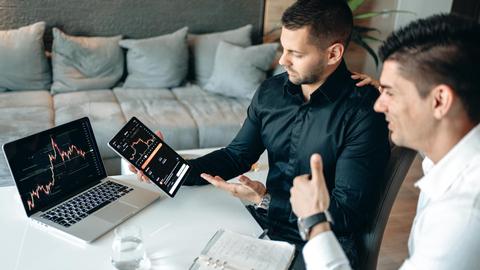 Two businessmen discuss digital trading insights with laptop and tablet in a modern office setting.