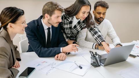 Diverse team of professionals working together on a laptop, analyzing charts and data in an office setting.