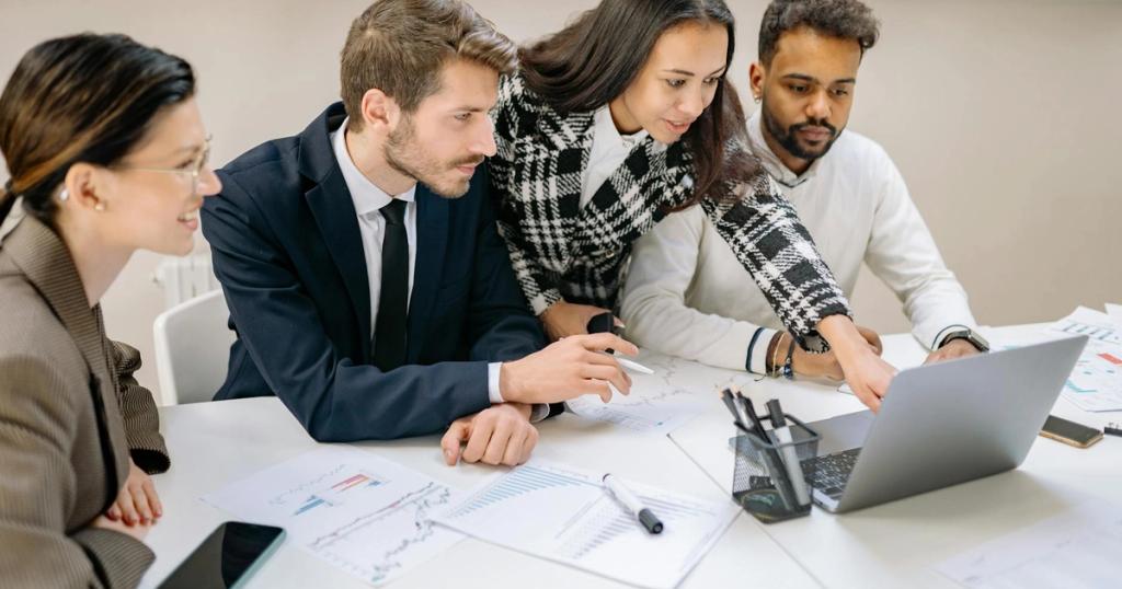 Diverse team of professionals working together on a laptop, analyzing charts and data in an office setting.