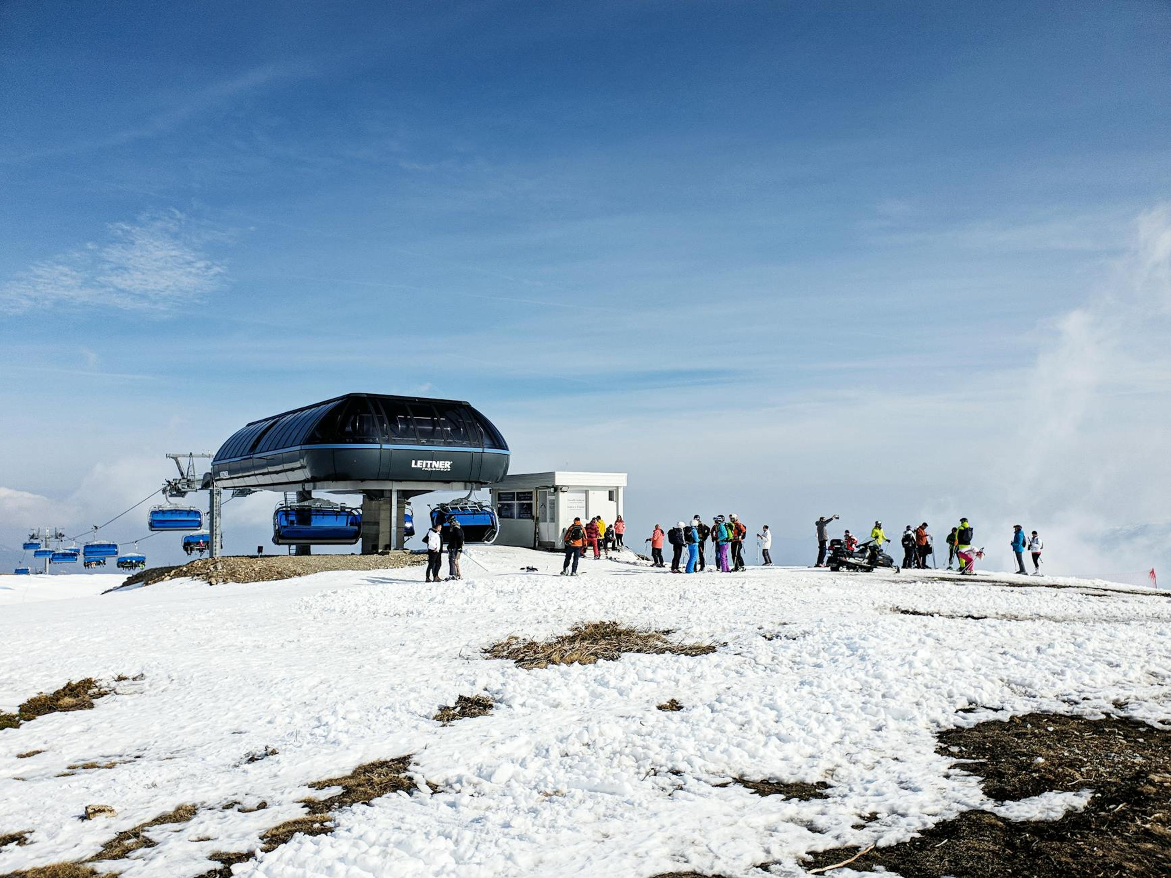 A group of skiers preparing near a modern chairlift station on a snowy mountain under a clear blue sky.