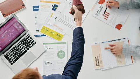 Business professionals engaging in a handshake over a desk filled with financial documents and a laptop.