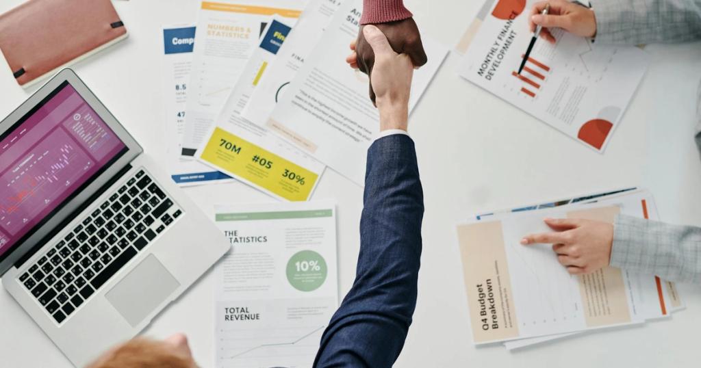Business professionals engaging in a handshake over a desk filled with financial documents and a laptop.