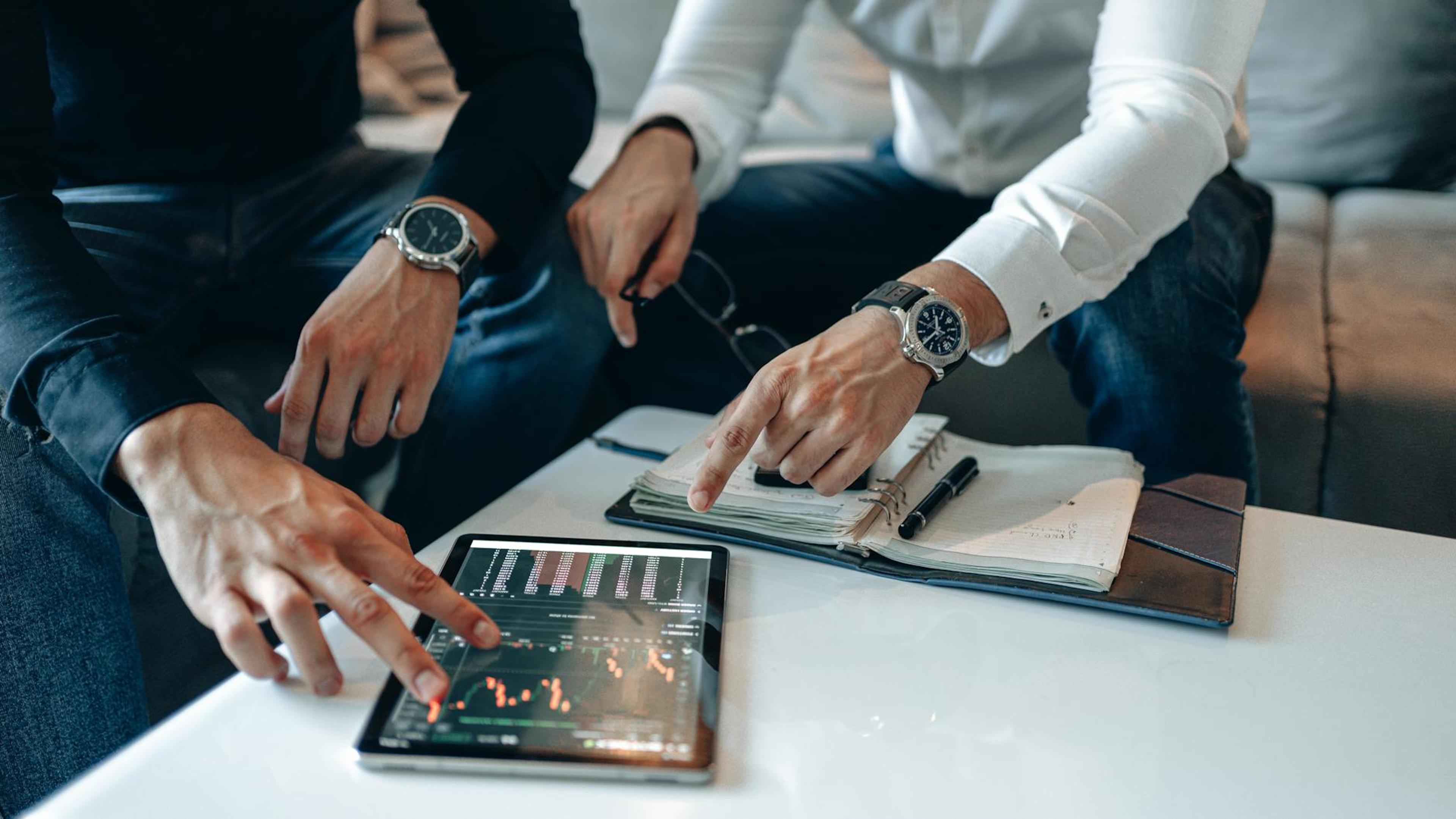 Two businessmen pointing at stock market data on a tablet, engaged in discussion.