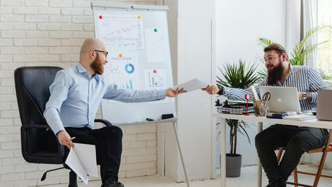Two business professionals exchanging documents in a modern office setting with charts and graphs.