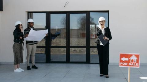 Real estate professionals with hard hats inspect a property for sale. Outdoor meeting with blueprints.
