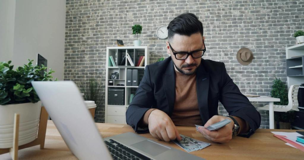 Businessman counting cash at a modern office desk with a laptop, showcasing finance management.