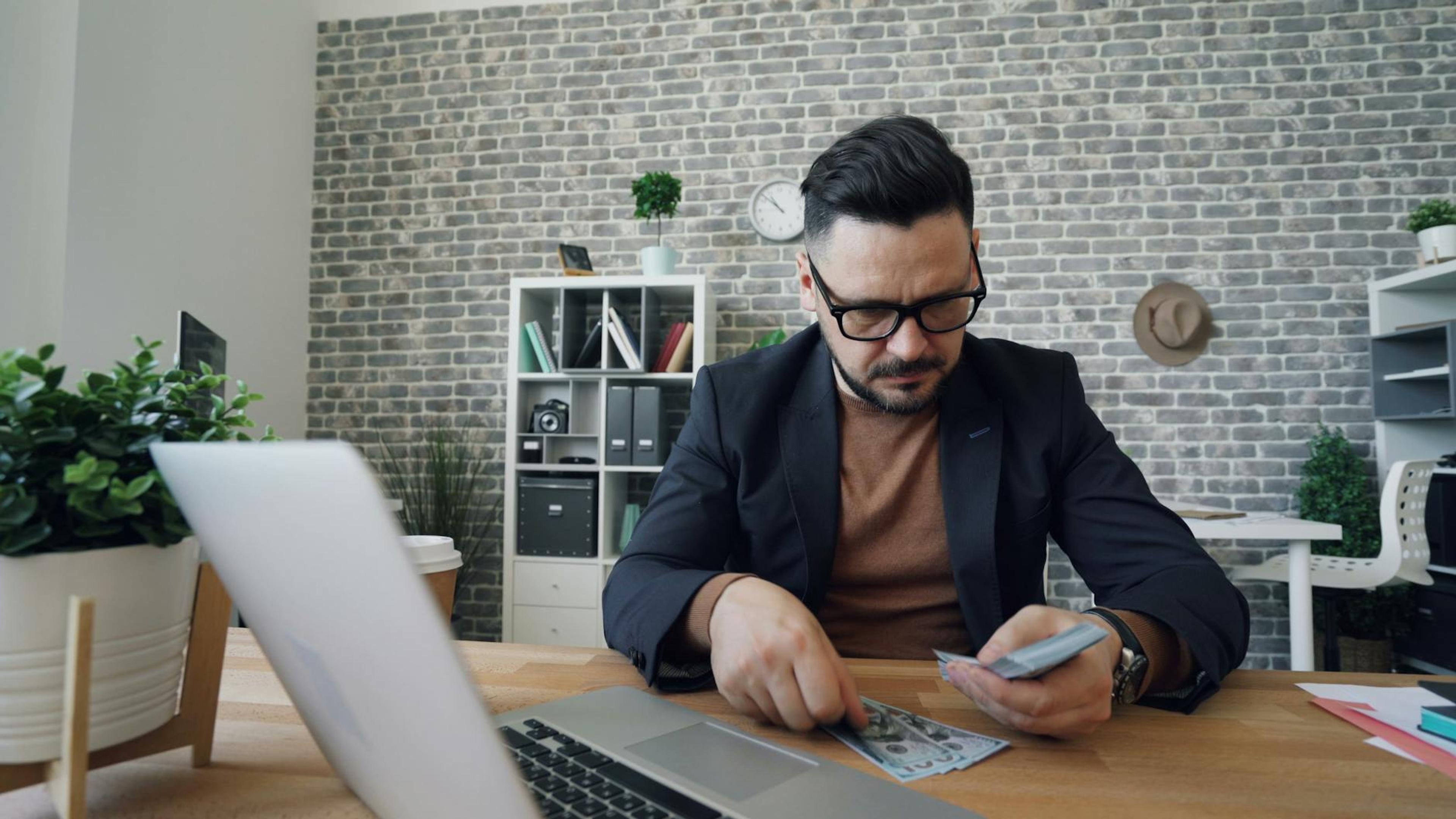 Businessman counting cash at a modern office desk with a laptop, showcasing finance management.