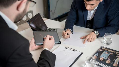 Two businessmen analyzing cryptocurrency market trends using a tablet and laptop in an office.