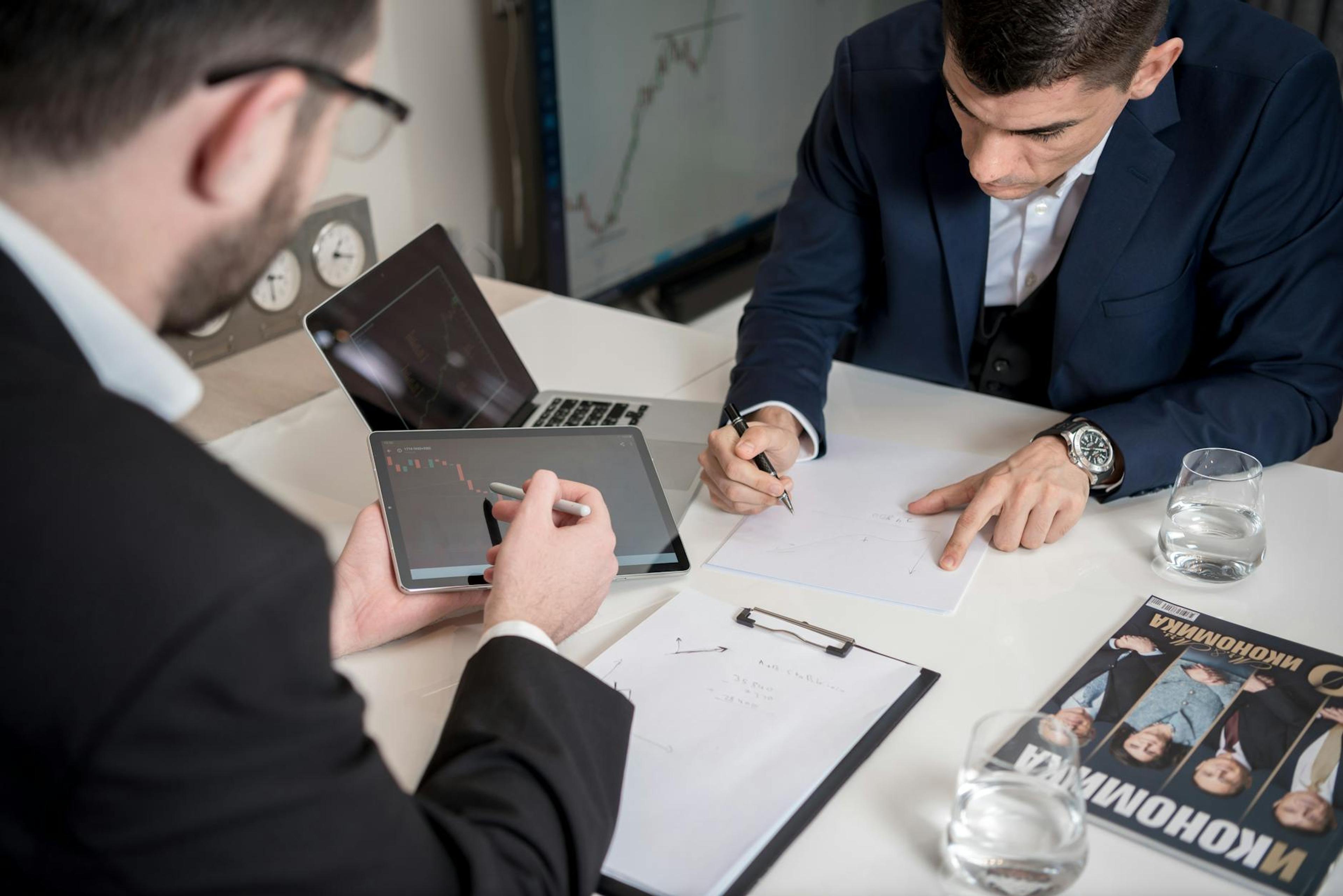 Two businessmen analyzing cryptocurrency market trends using a tablet and laptop in an office.
