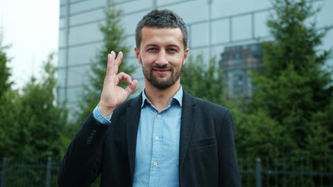 Businessman in suit giving an OK hand gesture outside modern building.