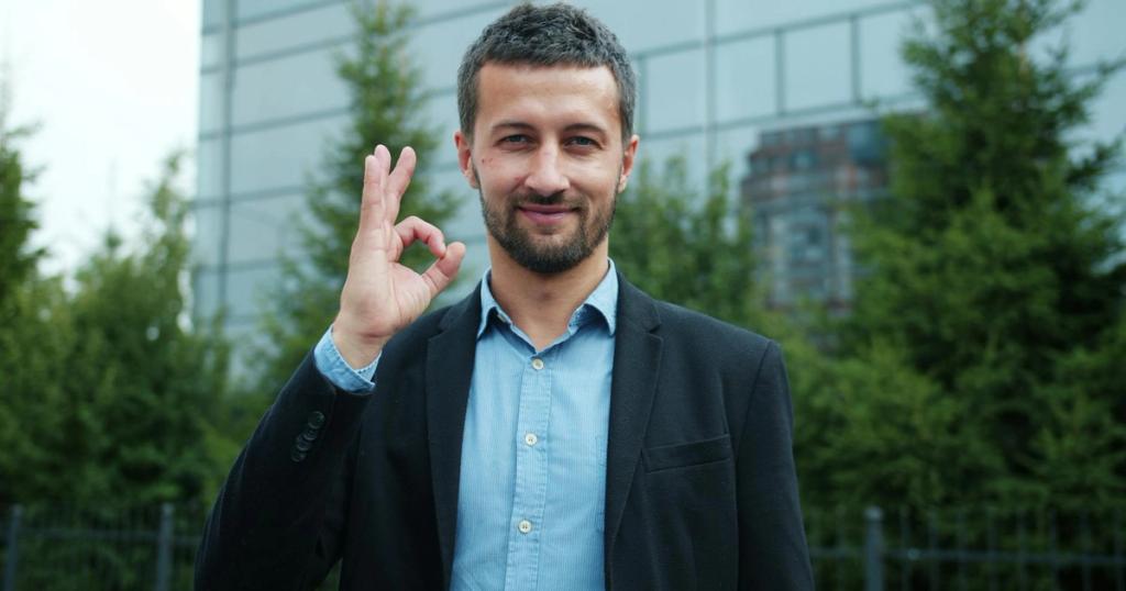 Businessman in suit giving an OK hand gesture outside modern building.