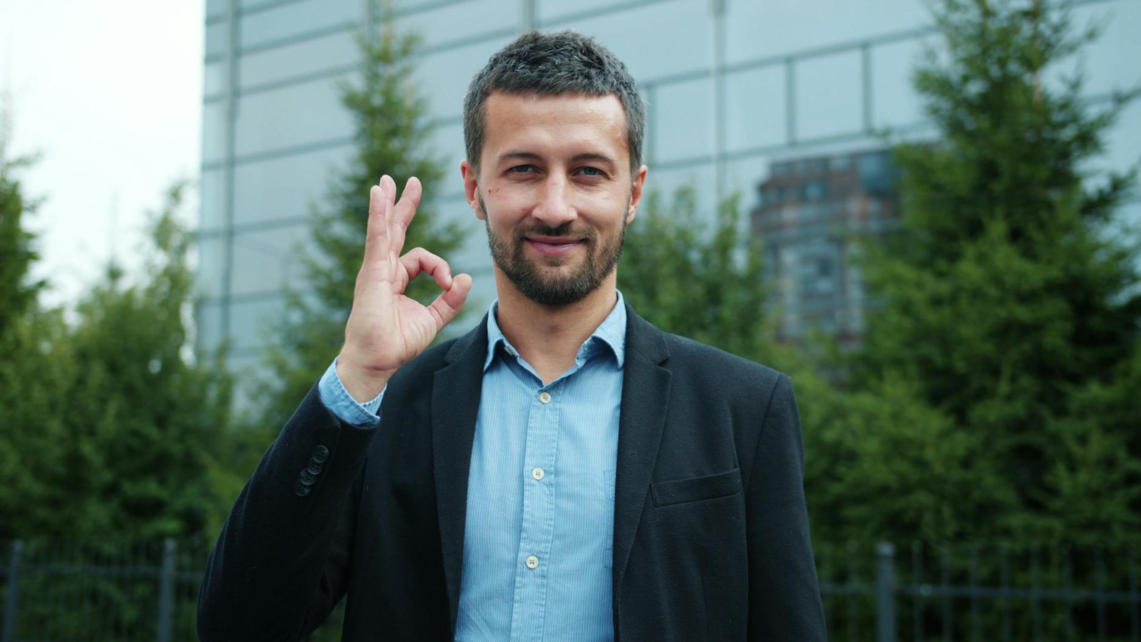 Businessman in suit giving an OK hand gesture outside modern building.