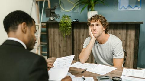 Man in office, appearing stressed during paperwork discussion, sitting opposite professional.