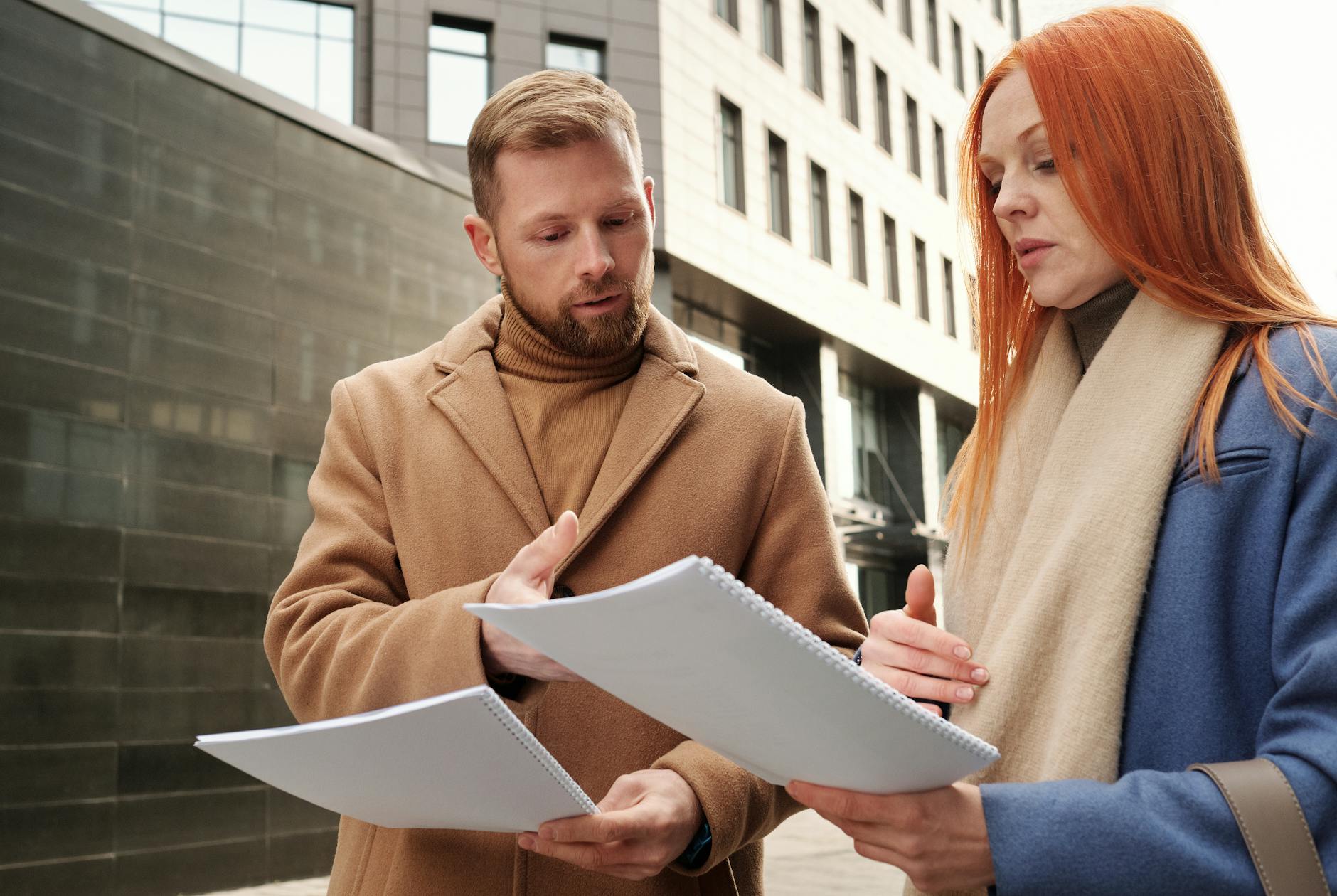 Two business professionals in coats discuss documents in an urban setting.