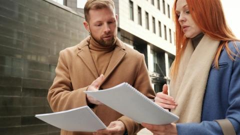 Two business professionals in coats discuss documents in an urban setting.