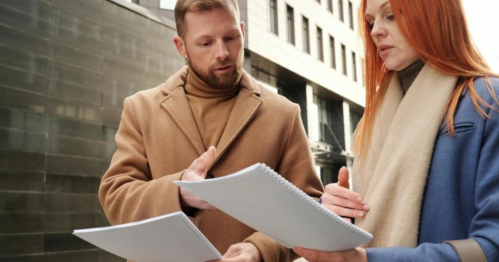 Two business professionals in coats discuss documents in an urban setting.