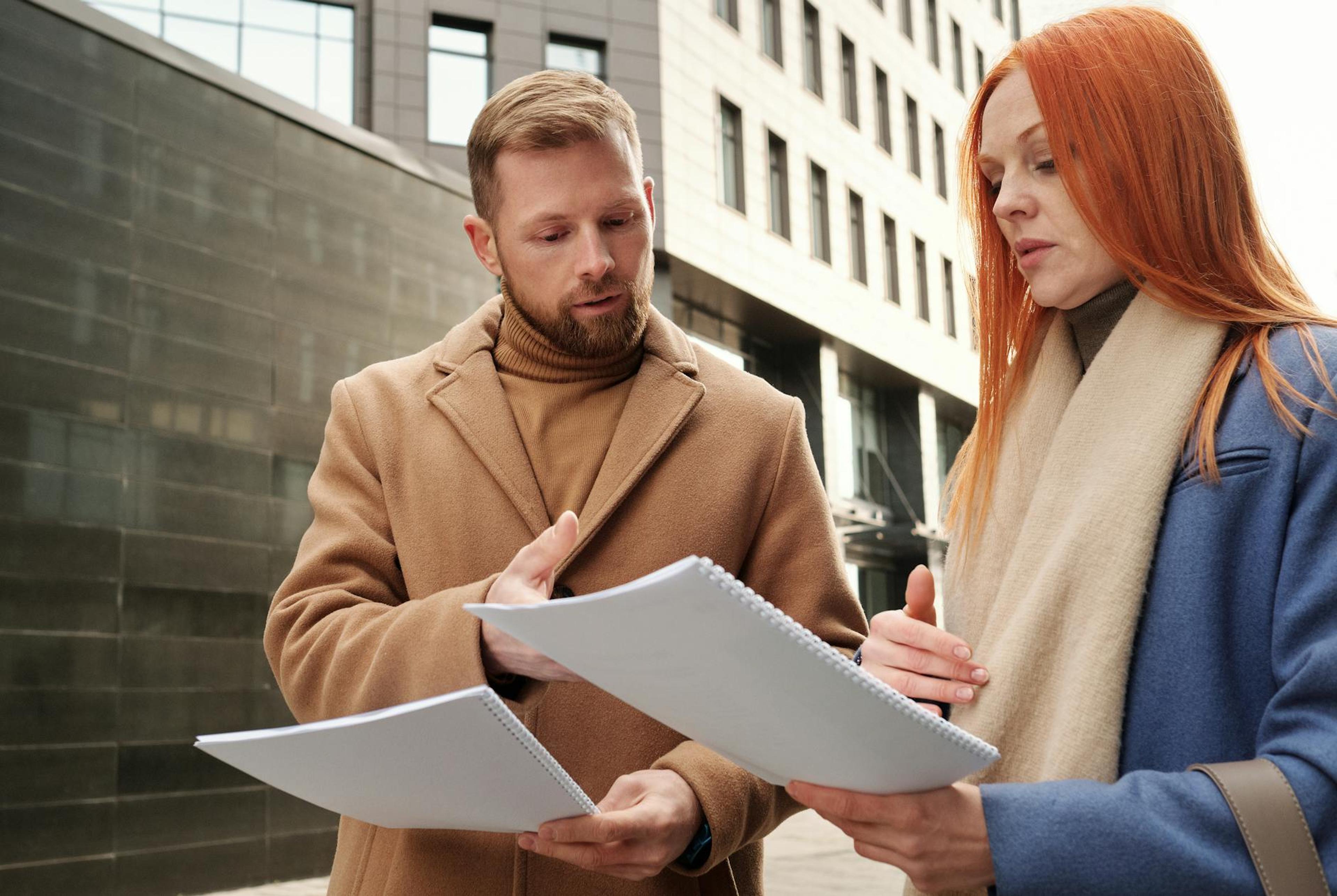 Two business professionals in coats discuss documents in an urban setting.