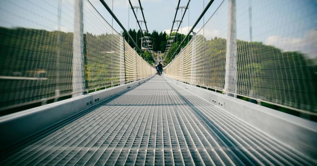 Long suspension bridge with people walking, showcasing modern architecture in a lush, sunny setting.