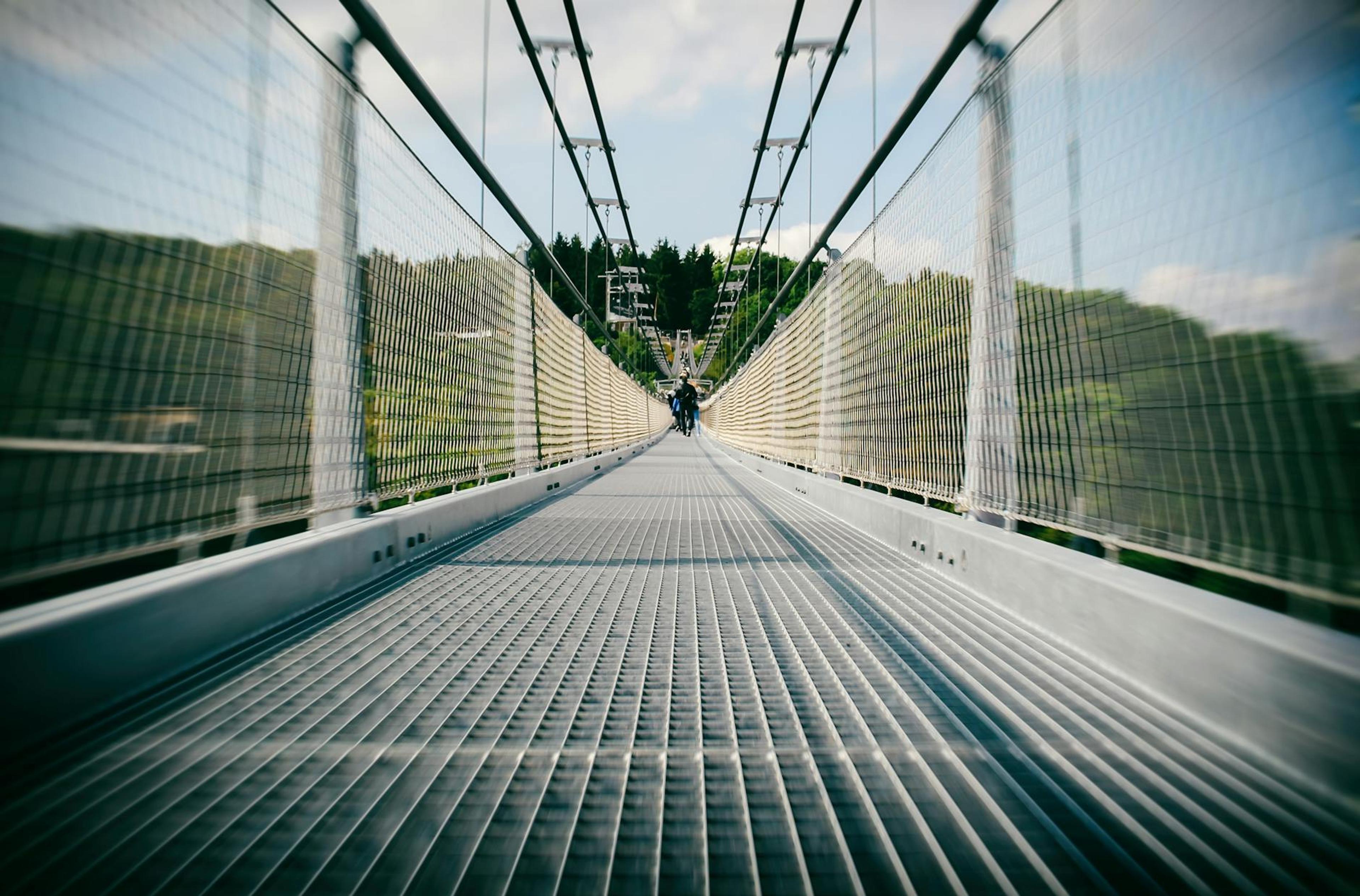 Long suspension bridge with people walking, showcasing modern architecture in a lush, sunny setting.