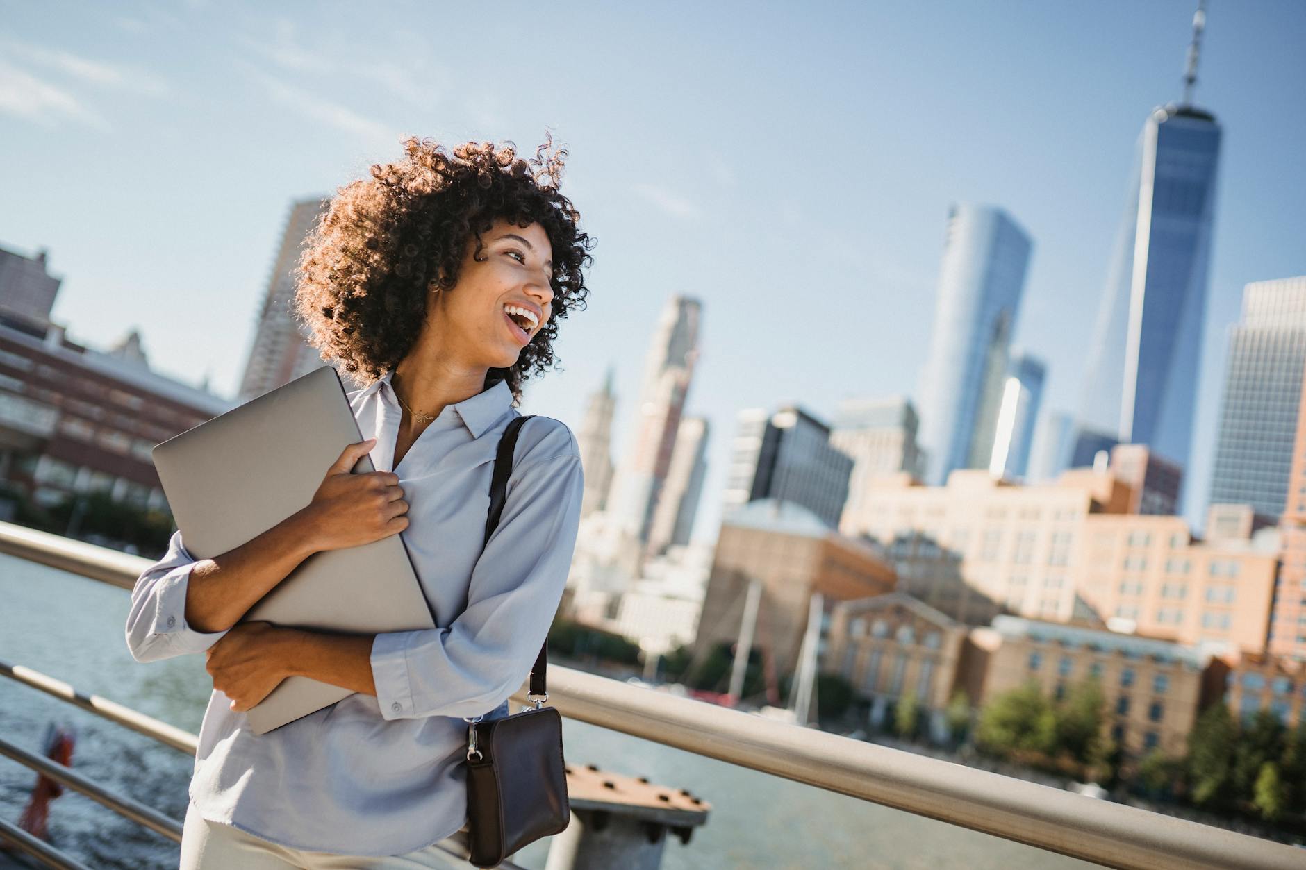 A happy young woman with a laptop enjoys the view of a city skyline by the river.