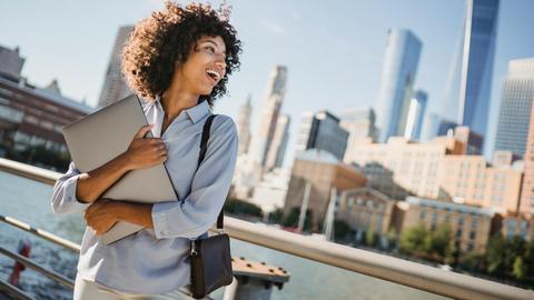 A happy young woman with a laptop enjoys the view of a city skyline by the river.