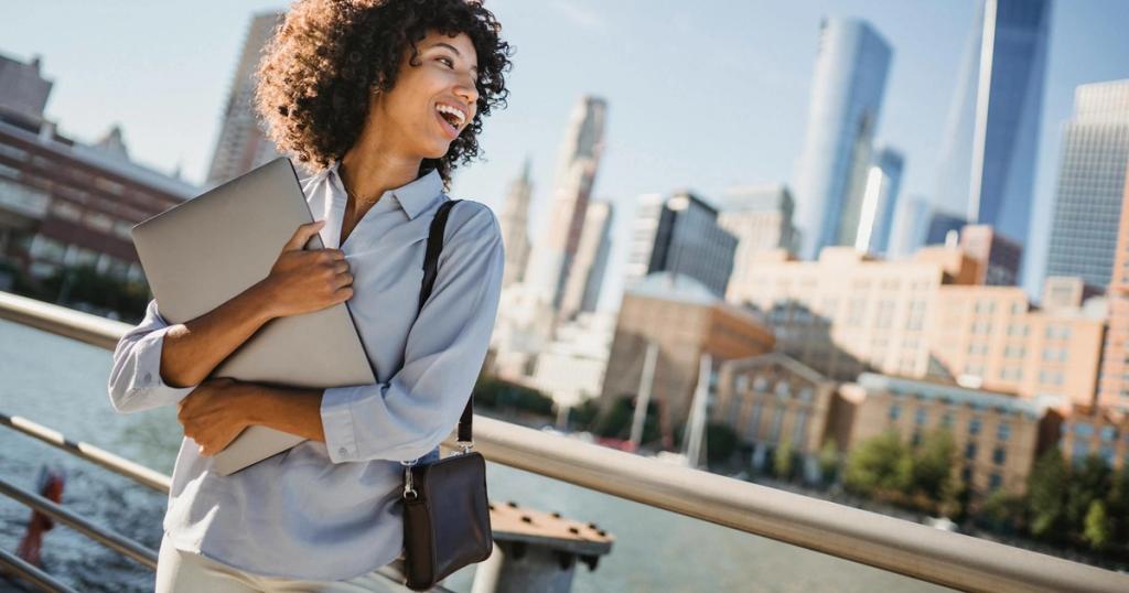 A happy young woman with a laptop enjoys the view of a city skyline by the river.