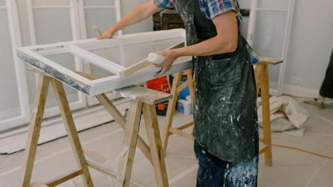 Unrecognizable male master wearing dirty apron standing near window with white frames on wooden workbench while working in professional studio