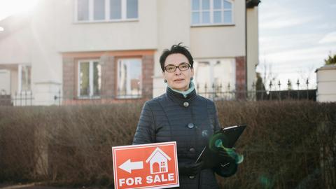 Woman realtor with eyeglasses holding a clipboard and 'For Sale' sign outside a house.