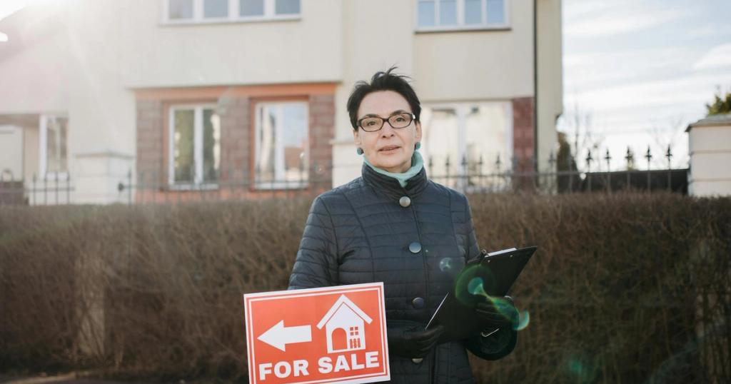 Woman realtor with eyeglasses holding a clipboard and 'For Sale' sign outside a house.