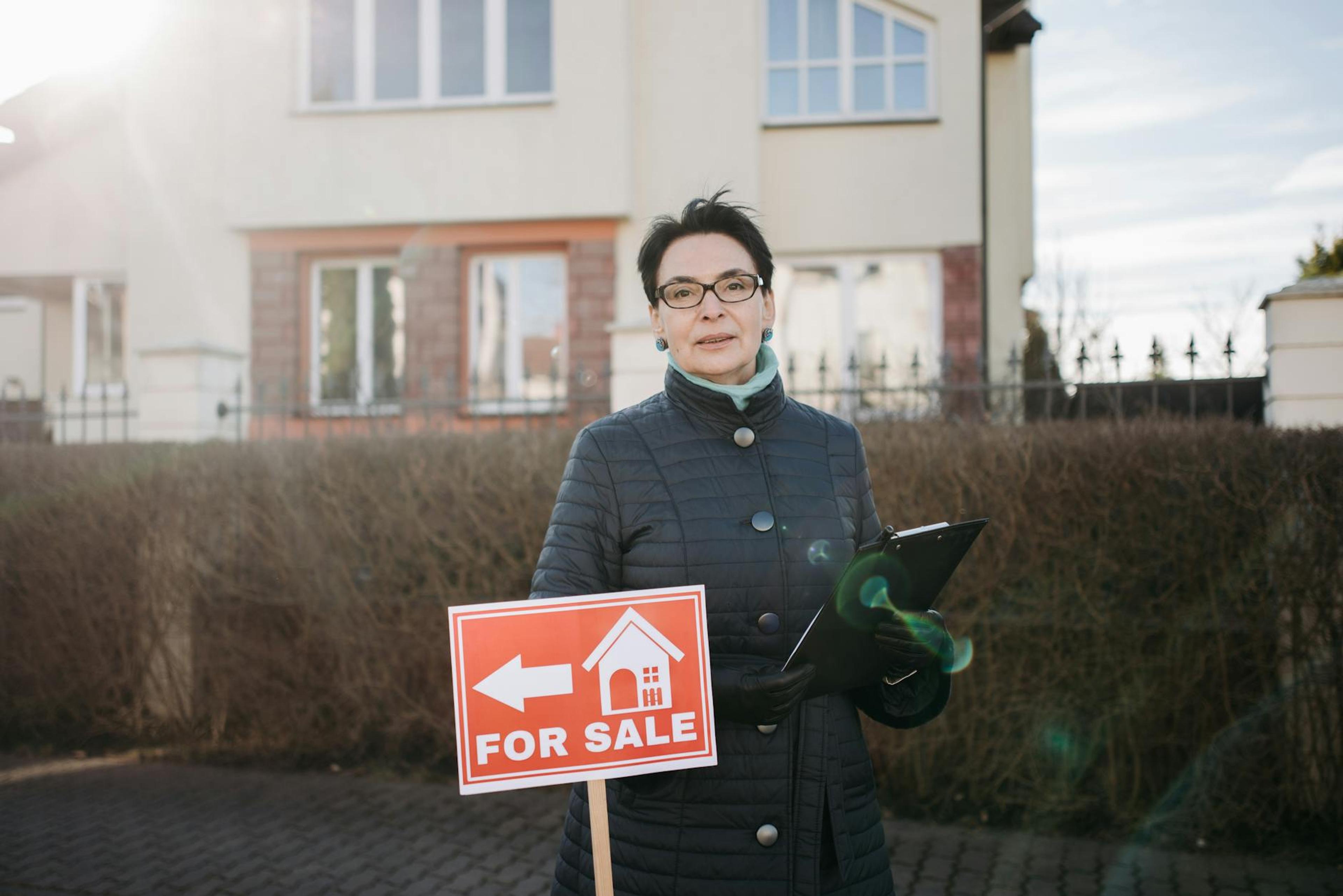 Woman realtor with eyeglasses holding a clipboard and 'For Sale' sign outside a house.