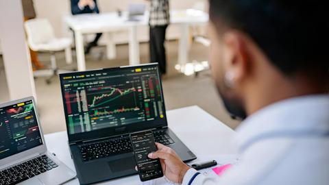 A businessman using smartphone and laptop to track stock market trends in a modern office setting.