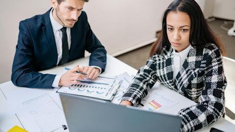A man and woman collaborate on business analysis at a desk with charts in an office setting.
