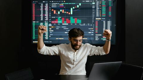 Businessman celebrates stock market success with hands raised in excitement at a trading desk.