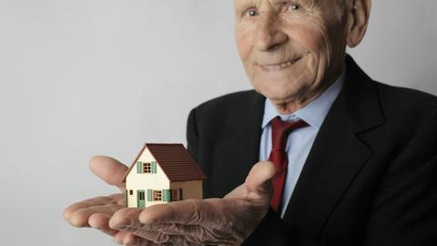 Elderly man in business attire holding a small house model, symbolizing real estate investment.