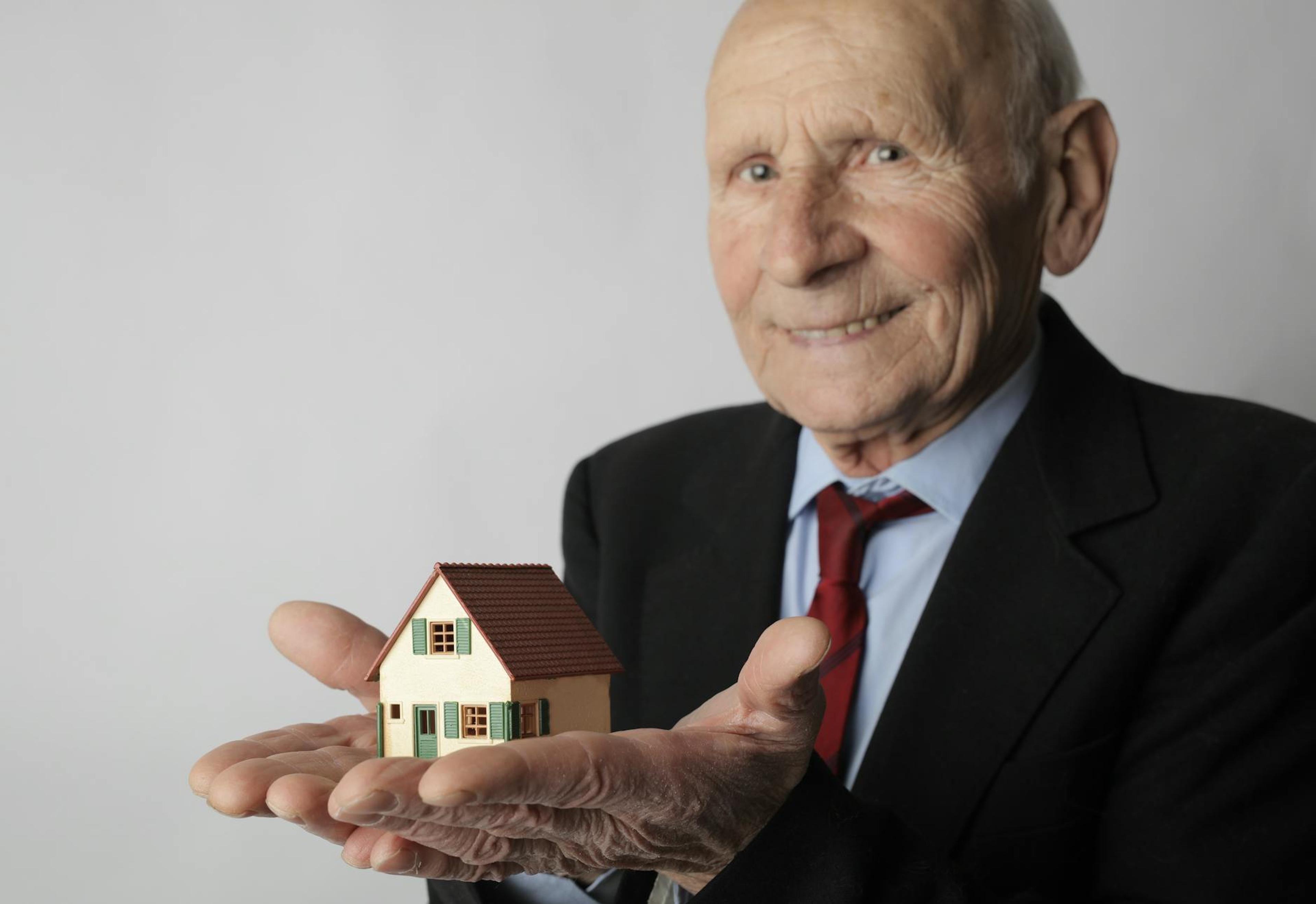 Elderly man in business attire holding a small house model, symbolizing real estate investment.