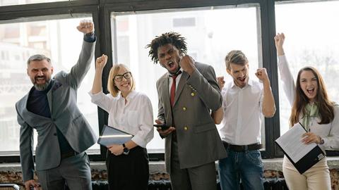 Group of diverse professionals celebrating achievement in office setting with enthusiasm.