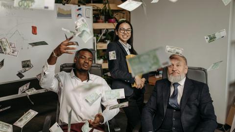 Diverse business team celebrating success with cash raining indoors in office.