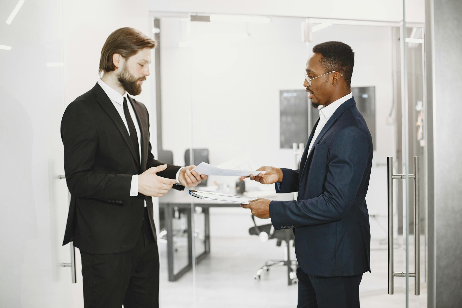 Two businessmen in formal suits exchanging documents in a modern office setting, symbolizing collaboration and partnership.