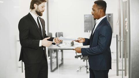 Two businessmen in formal suits exchanging documents in a modern office setting, symbolizing collaboration and partnership.