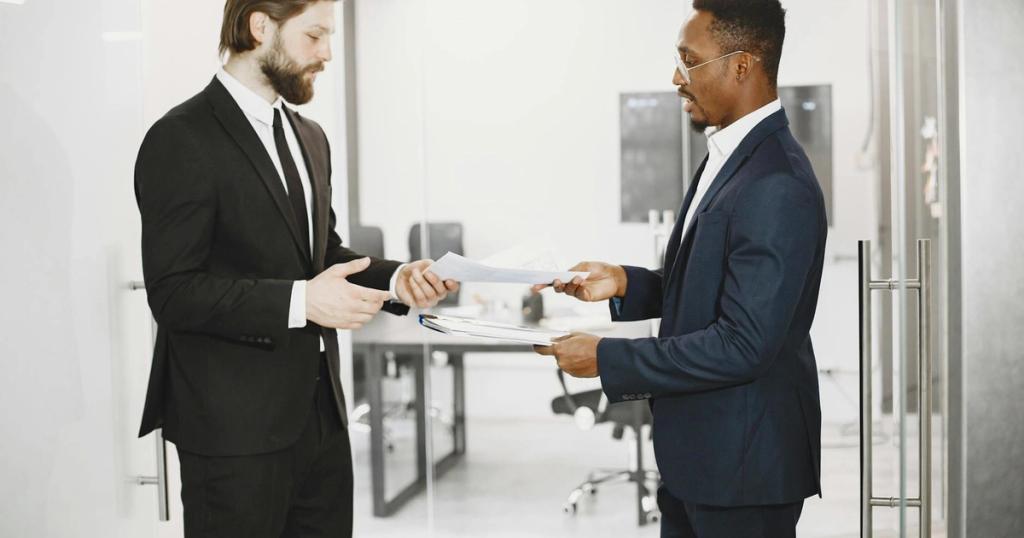 Two businessmen in formal suits exchanging documents in a modern office setting, symbolizing collaboration and partnership.