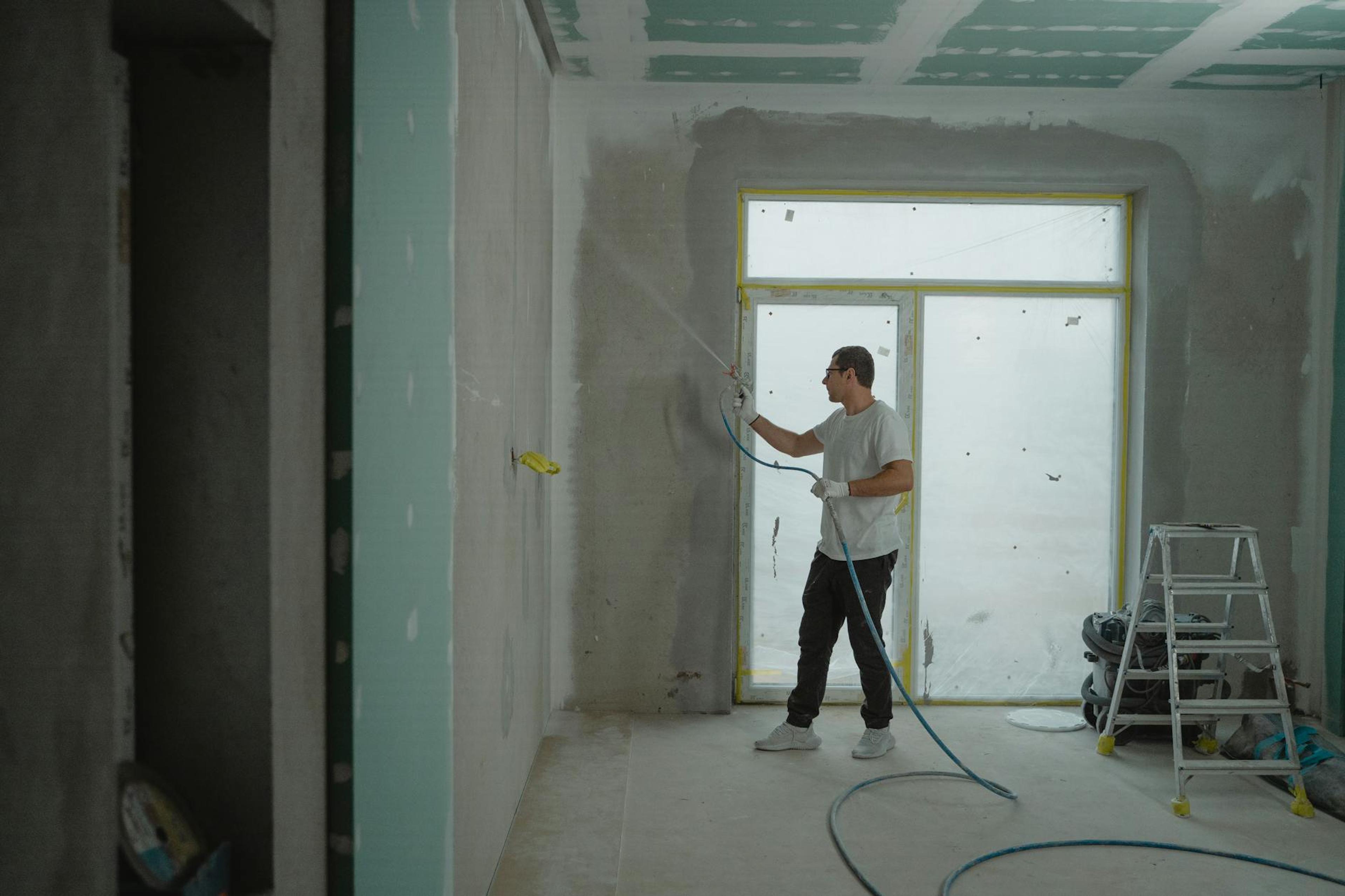 A man sprays paint on a wall while renovating an indoor room with professional equipment.