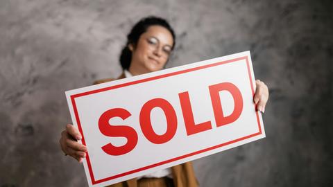 A smiling woman with eyeglasses holding a 'Sold' sign, signifying success in real estate sales.