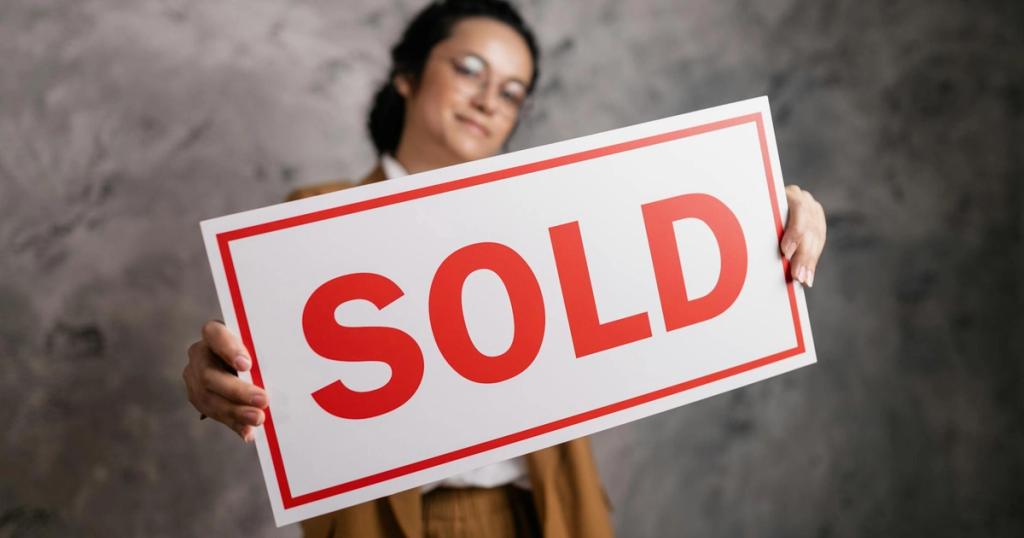 A smiling woman with eyeglasses holding a 'Sold' sign, signifying success in real estate sales.