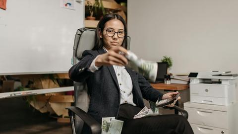 Confident businesswoman in eyeglasses counting dollar bills while sitting in a modern office.