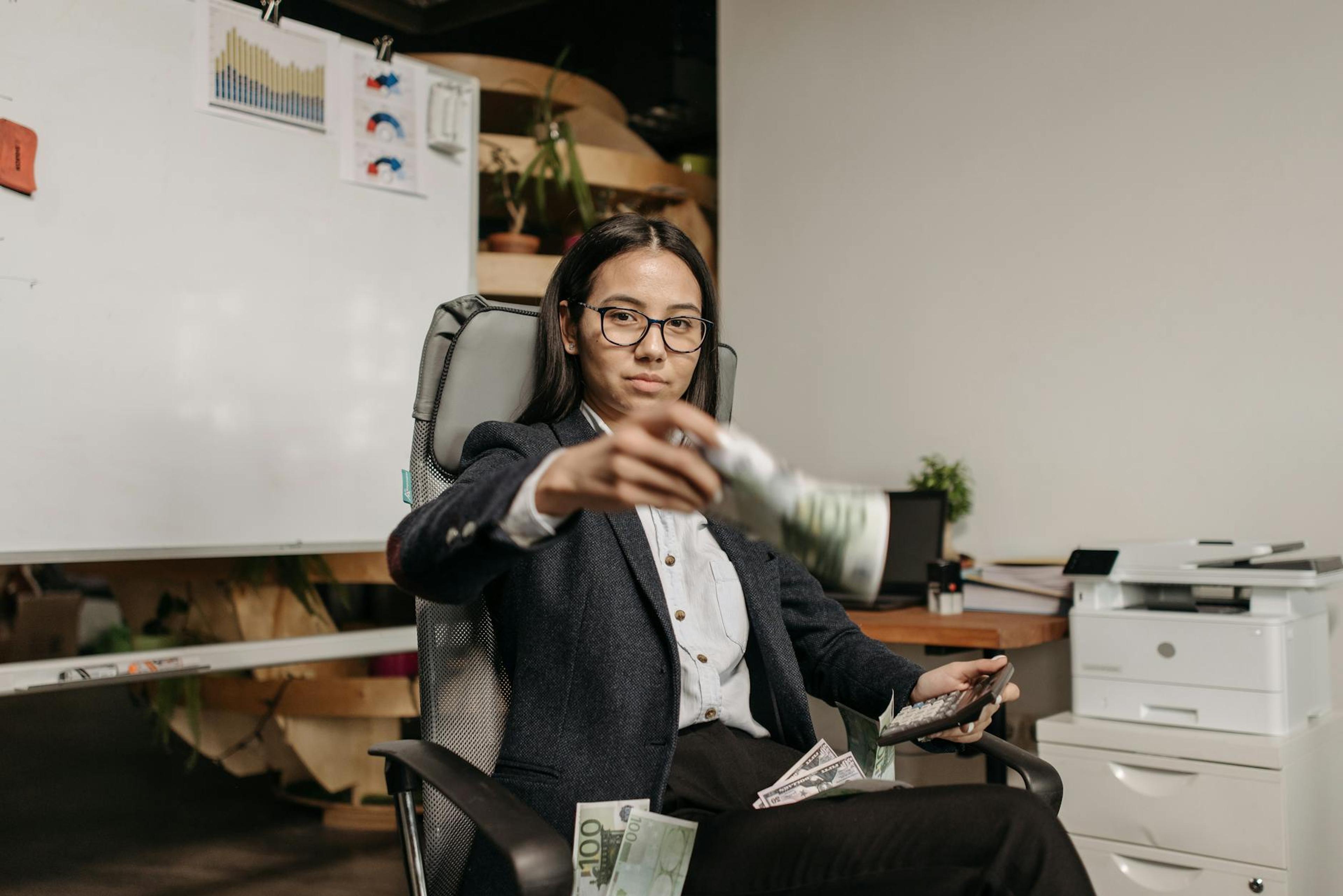 Confident businesswoman in eyeglasses counting dollar bills while sitting in a modern office.