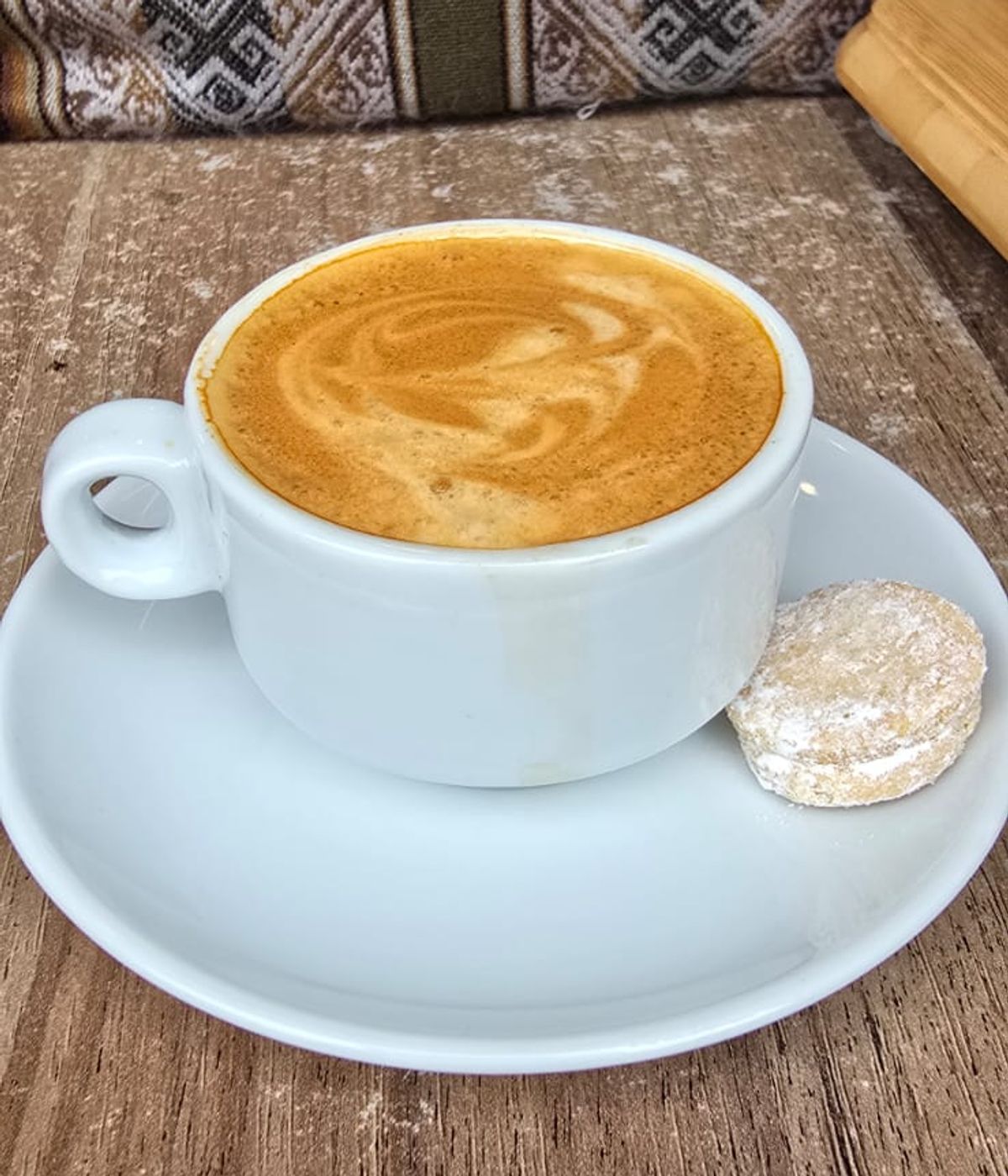 Taza de espresso con un mini alfajor, sobre mesa de madera y textil ayacuchano