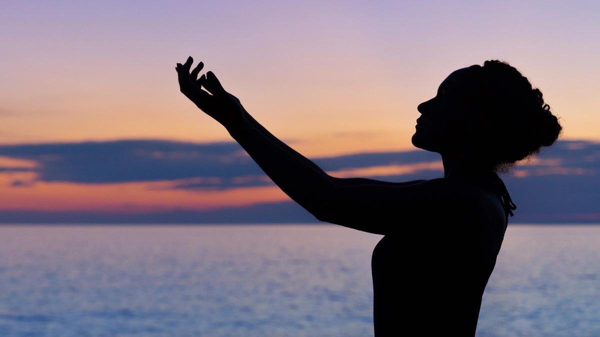 The silhouette of a woman reaching her arms in front of her with a pink and blue sky after sunset behind her.