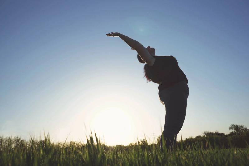 An average-bodied woman stands outside on grass and gently bends backwards.
