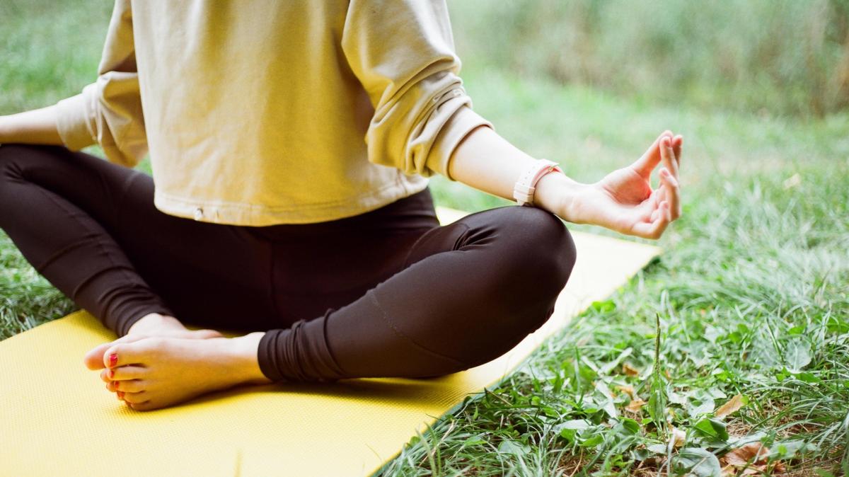 A person wearing a yellow sweatshirt sits with their feet together on a yellow yoga mat on grass.